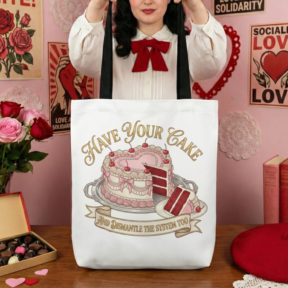 A person holds a tote bag with a pink cake illustration and text "Have Your Cake and Dismantle the System Too." The room is decorated with red and pink accents, posters, roses, and chocolates, creating a playful yet rebellious tone.