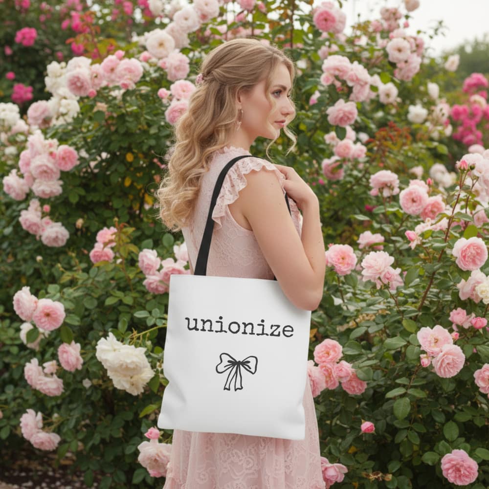 A woman in a pink dress stands in a rose garden. She holds a white tote bag with "unionize" and a bow design, conveying elegance and empowerment.