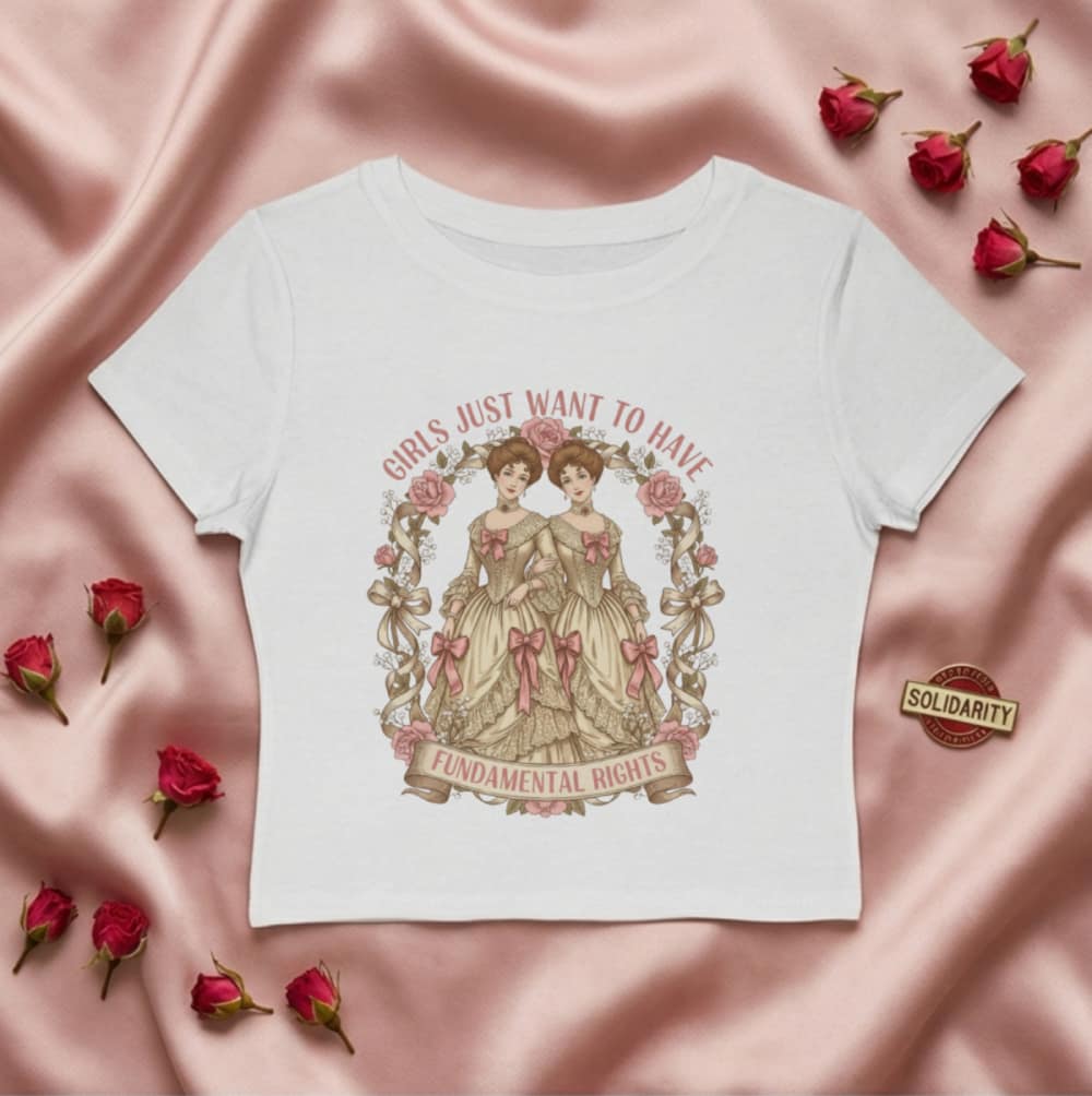 White crop top on pink satin with a vintage illustration of two girls in dresses. Text reads, "Girls Just Want to Have Fundamental Rights." Surrounded by roses.